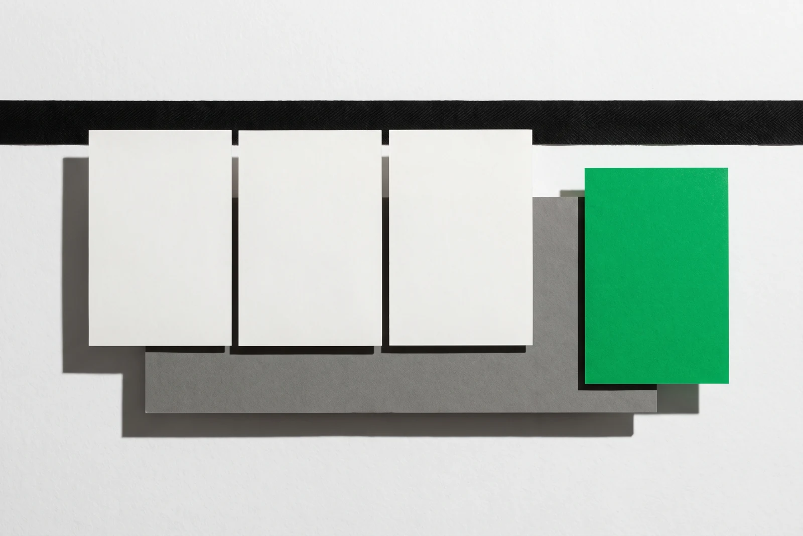 Trois cartes de papier vierge poussées jusqu’à buter contre une bande de toile noire, une quatrième carte verte restée en arrière, ombres dures projetées vers le bas à droite.