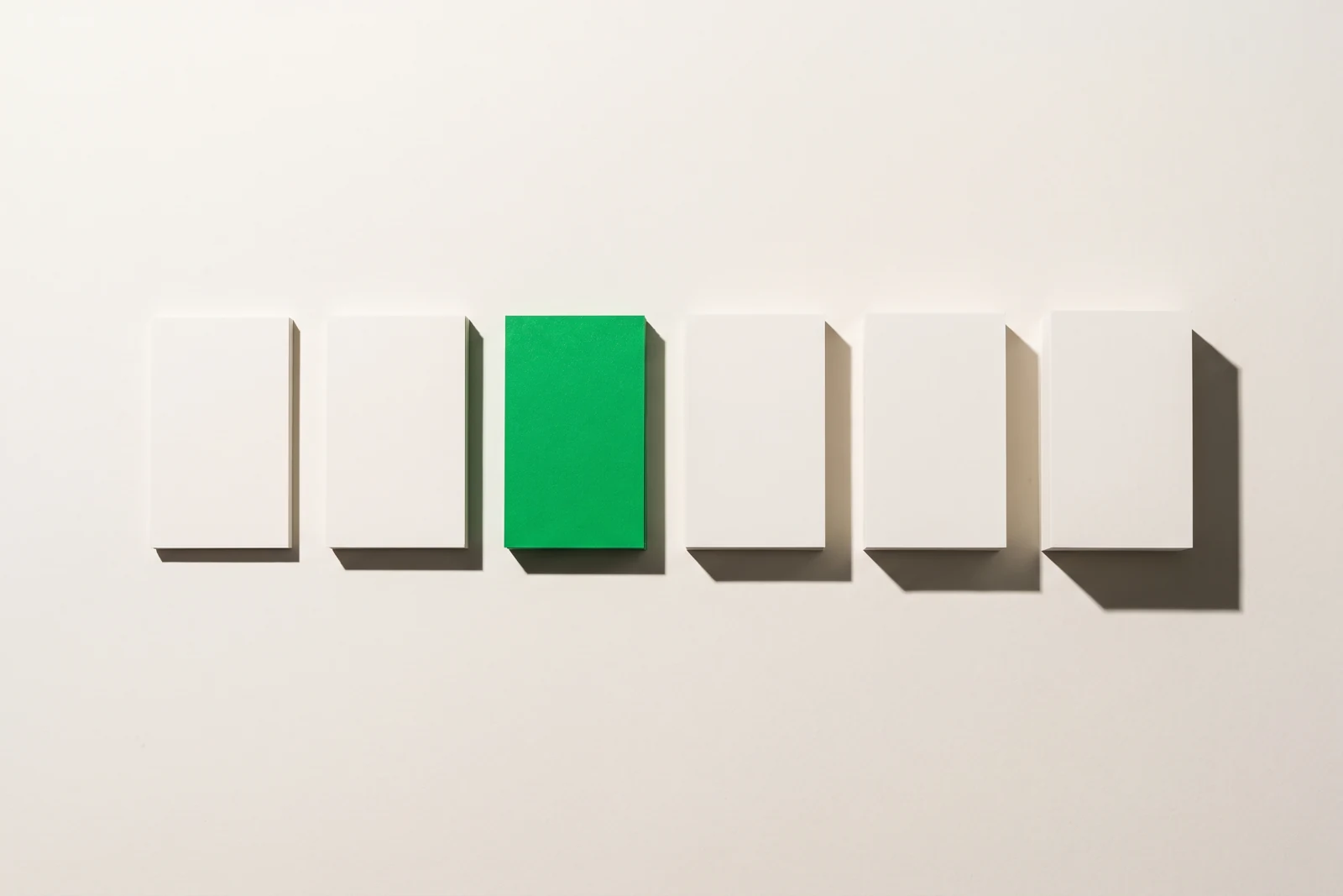Six piles de cartes de papier mat alignées sur la même base, de la plus fine à la plus épaisse, dont une pile coiffée d'une carte verte, ombres dures projetées dans une seule direction
